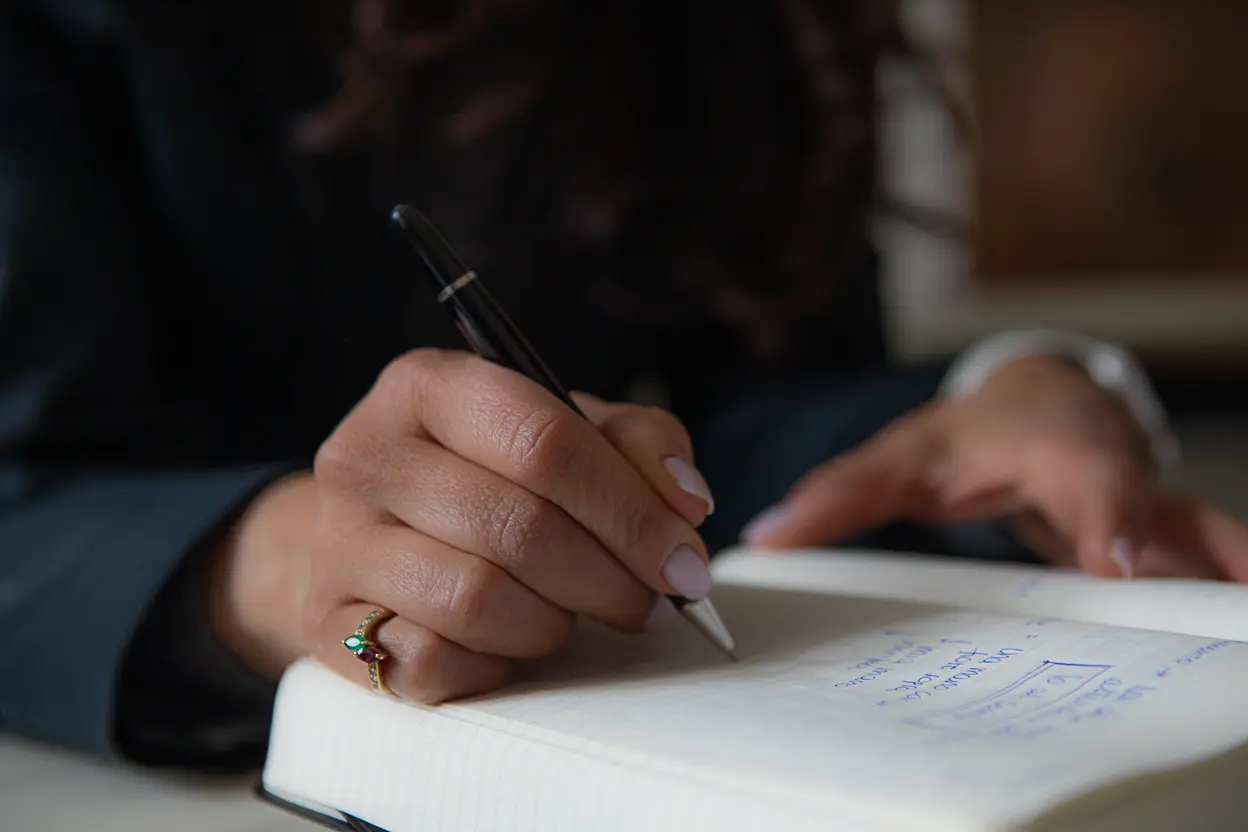 Mano di una persona che scrive su un taccuino con una penna nera, con un anello decorato sull'anulare e smalto chiaro sulle unghie.
