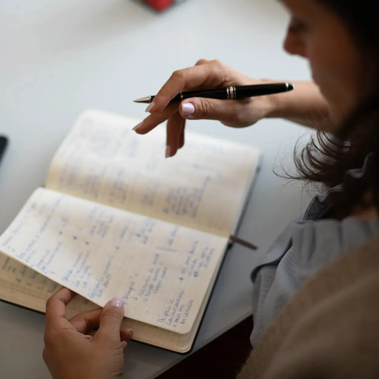 Primo piano di una persona che tiene una penna nera mentre legge o consulta un quaderno aperto con appunti scritti a mano.