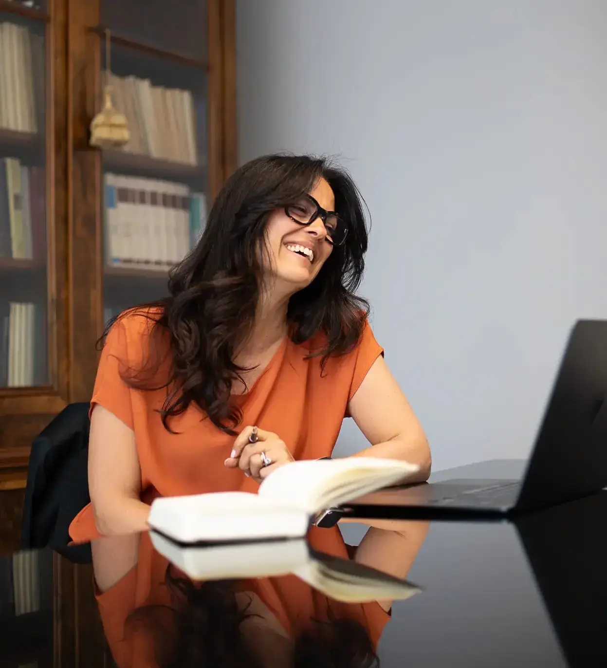 Donna che sorride mentre lavora a un tavolo davanti a un laptop con un libro aperto e una penna in mano, con una libreria sullo sfondo.