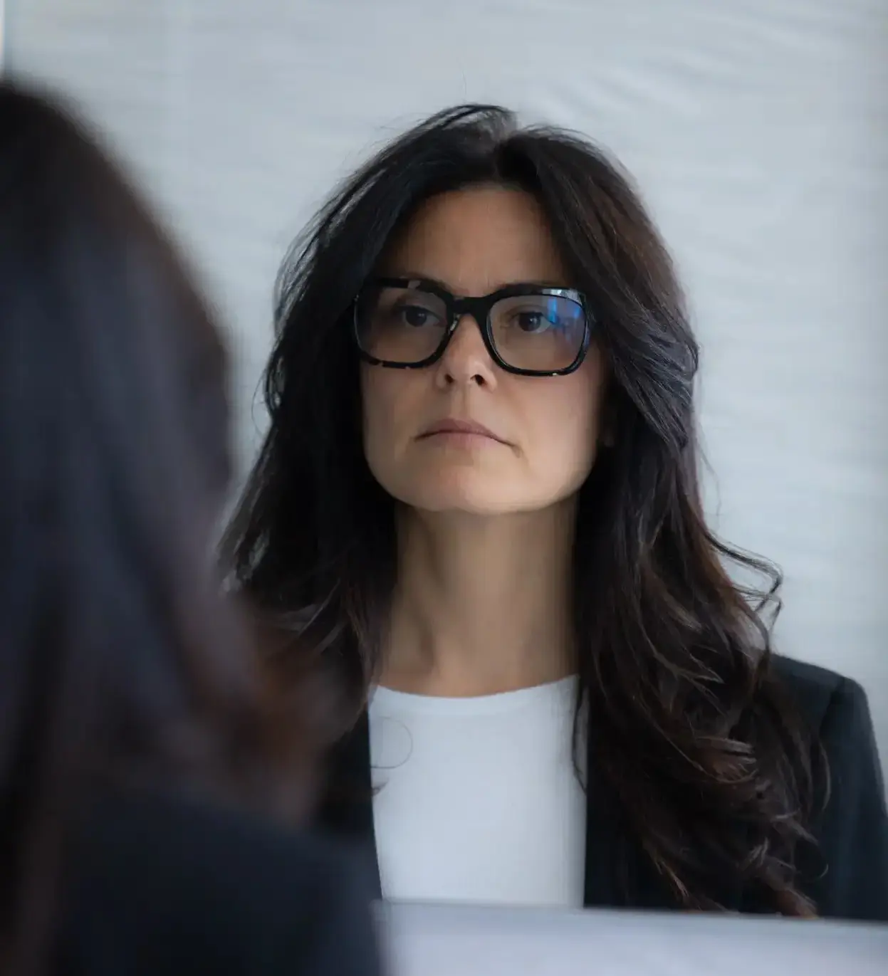 Donna con lunghi capelli scuri e occhiali neri che si guarda riflessa in uno specchio con una espressione seria, indossa una maglietta bianca e una giacca nera.