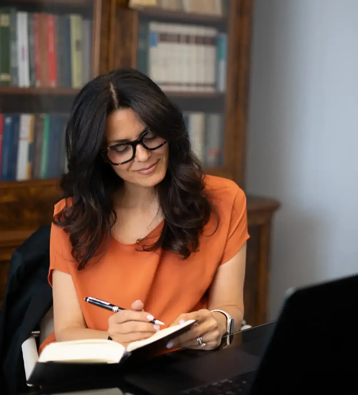 Donna con capelli castano scuro e occhiali, indossa una maglietta arancione mentre scrive su un taccuino con una penna, seduta davanti a un computer portatile con una libreria di libri sullo sfondo.