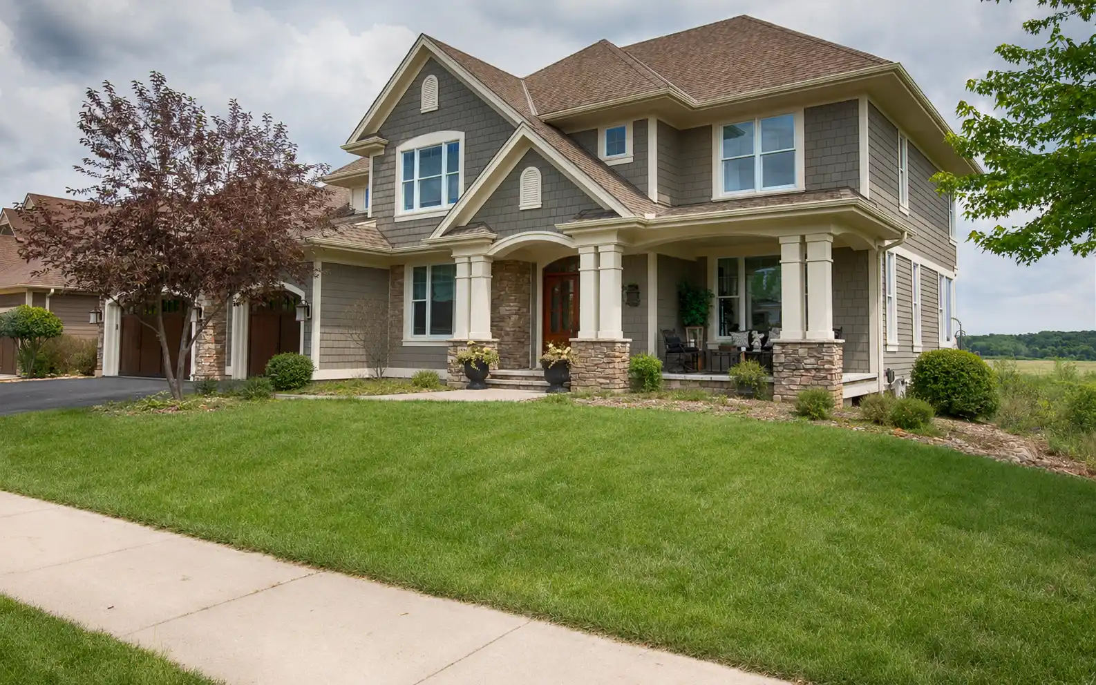 Two-story house with gray siding, stone accents, front porch, and a green lawn under a cloudy sky.