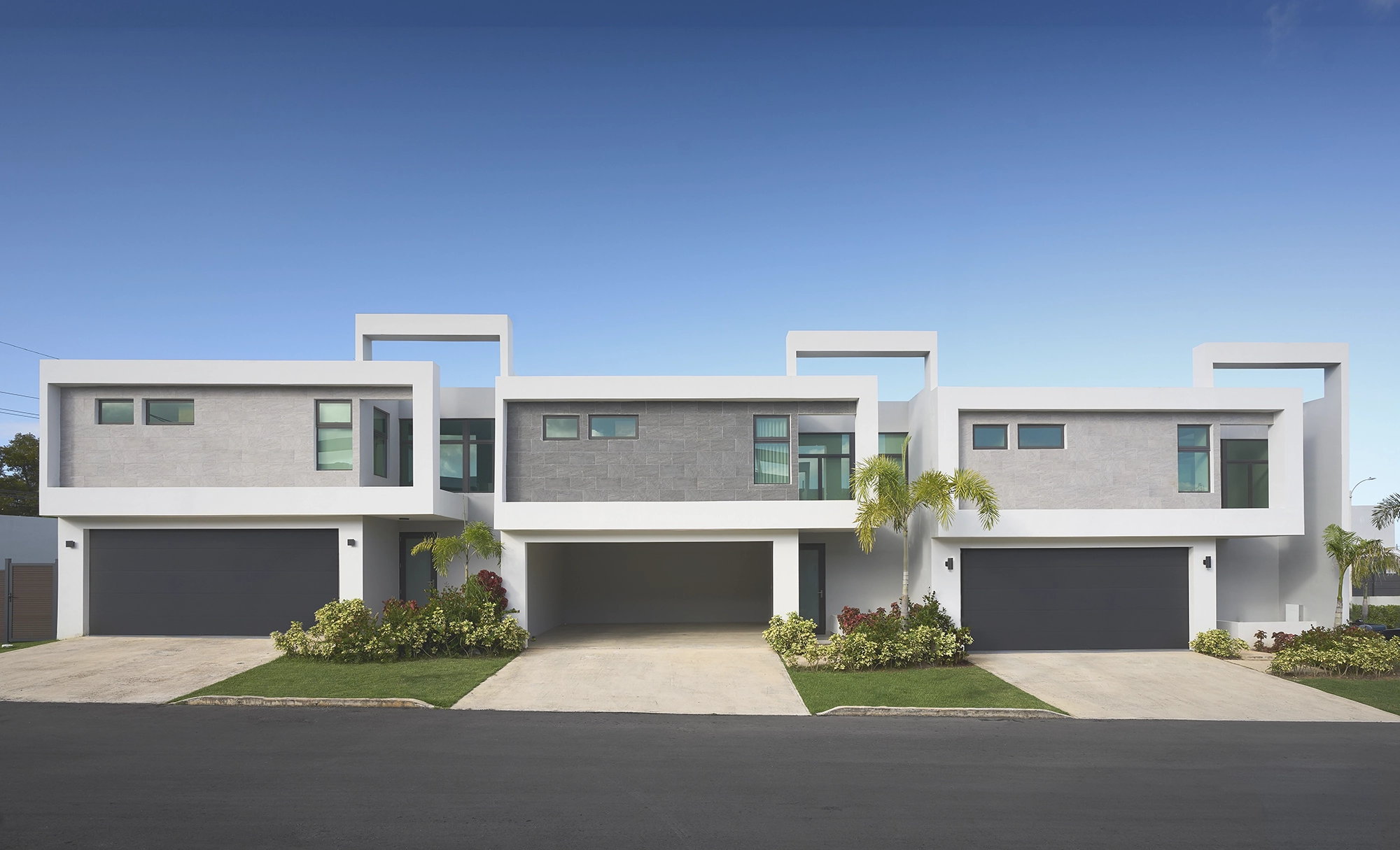 Moderno complejo de tres casas adosadas blancas con garajes y jardines delanteros bajo un cielo azul despejado.