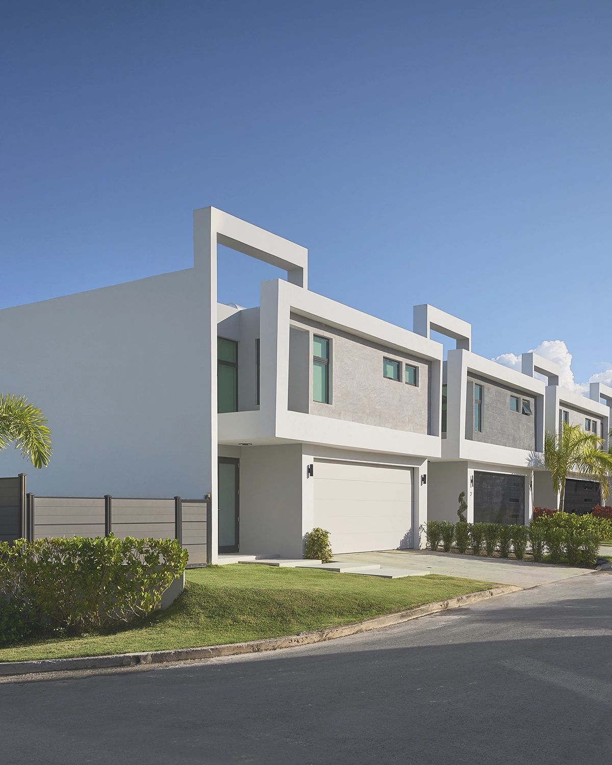 Hilera de modernas casas blancas de dos plantas con grandes ventanales, garajes y césped bien cuidado bajo un cielo azul.