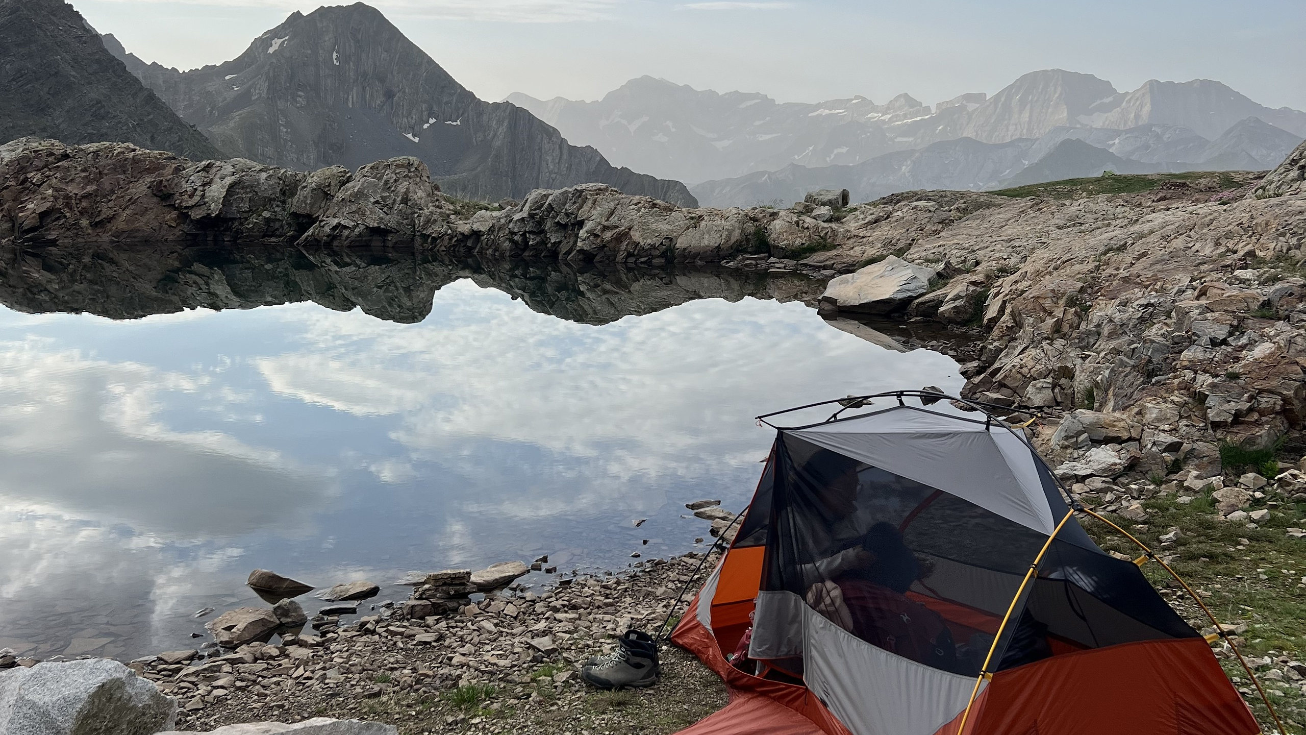 Tente de bivouac au bord d'un laquet de montagne, sommets et nuages reflétés dans l'eau