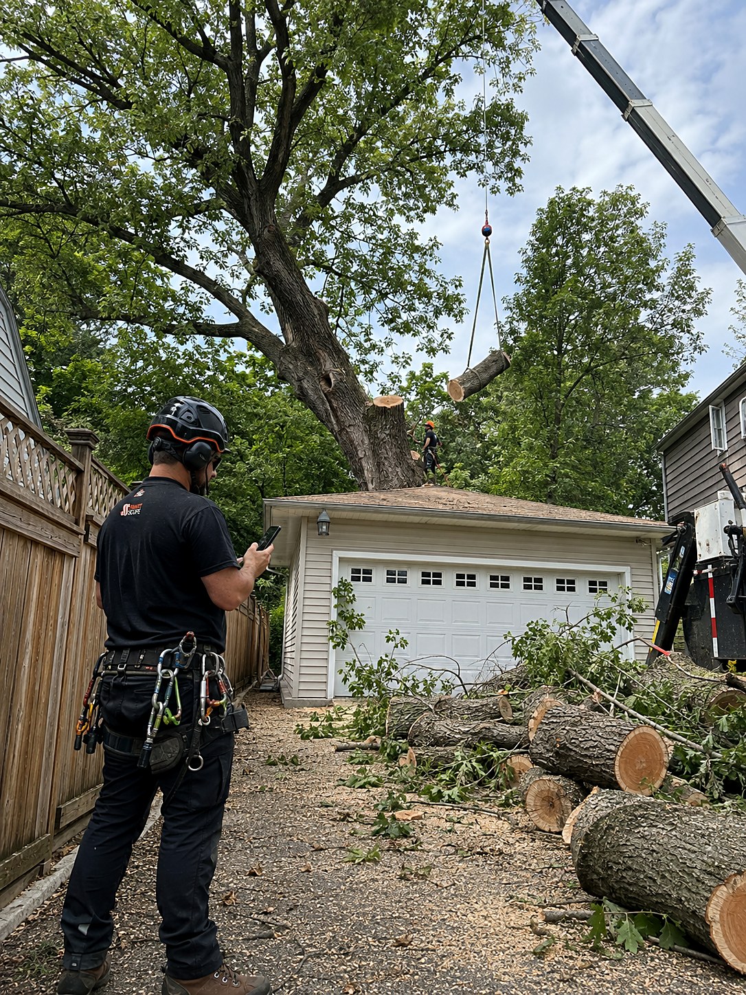 Crane lifting a log section off a large oak leaning over a garage while a Smart Scope arborist prices the removal on his phone in a tight fenced drop zone