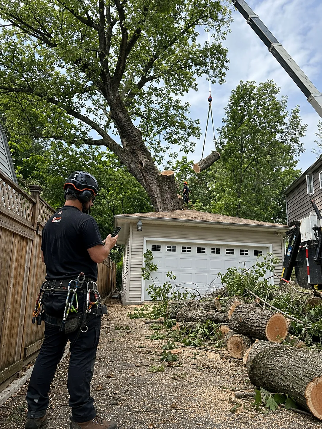 Crane lifting a log section off a large oak leaning over a garage while a Smart Scope arborist prices the removal on his phone in a tight fenced drop zone