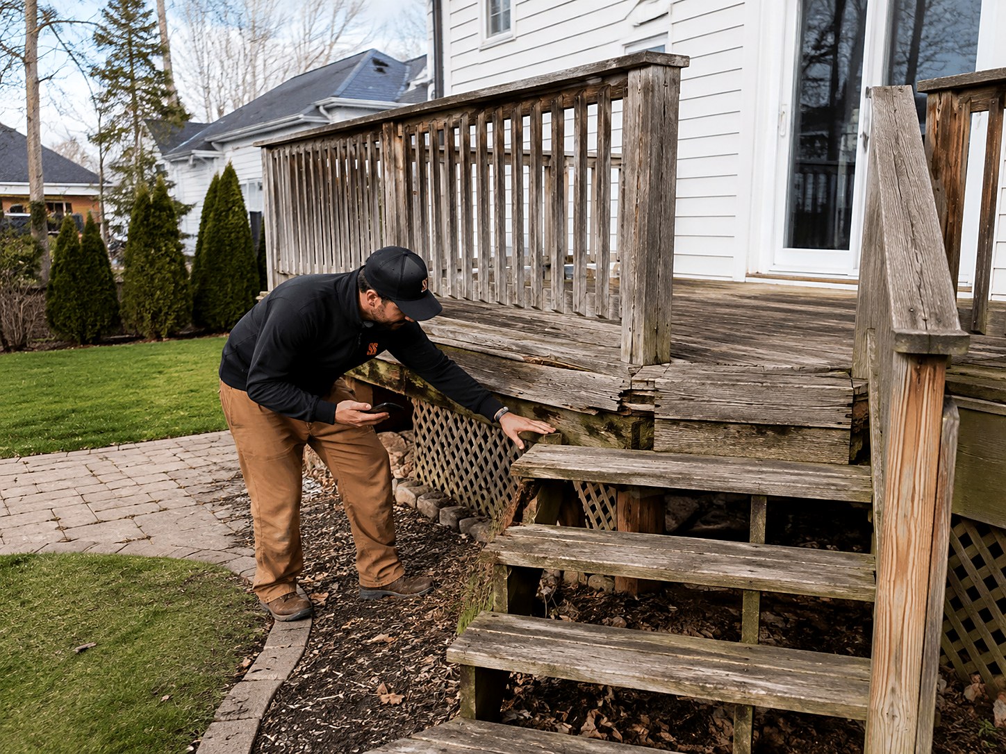 Contractor with a phone pressing on a soft rim joist while inspecting a weathered wood deck with worn railing and stairs