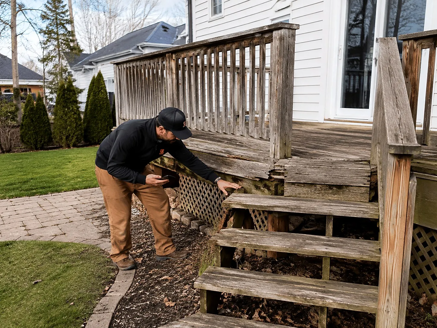 Contractor with a phone pressing on a soft rim joist while inspecting a weathered wood deck with worn railing and stairs