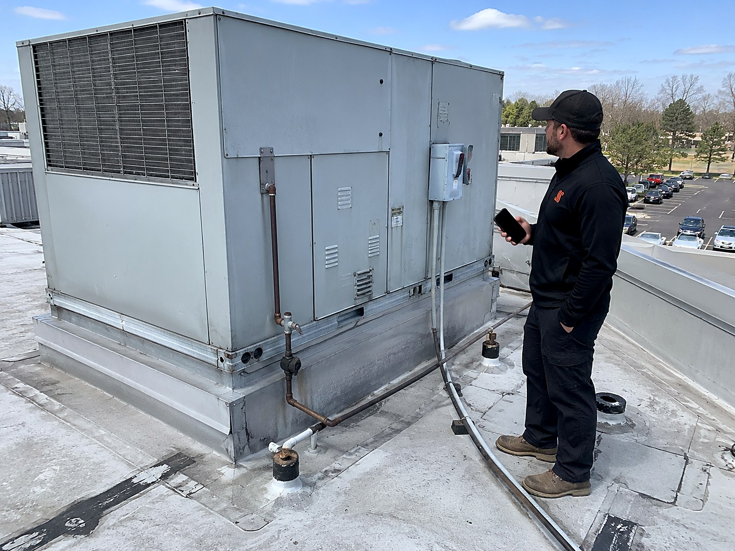 Mechanical contractor with a phone assessing an aging rooftop unit on its curb, with gas piping and an electrical disconnect on the side and the parking lot below