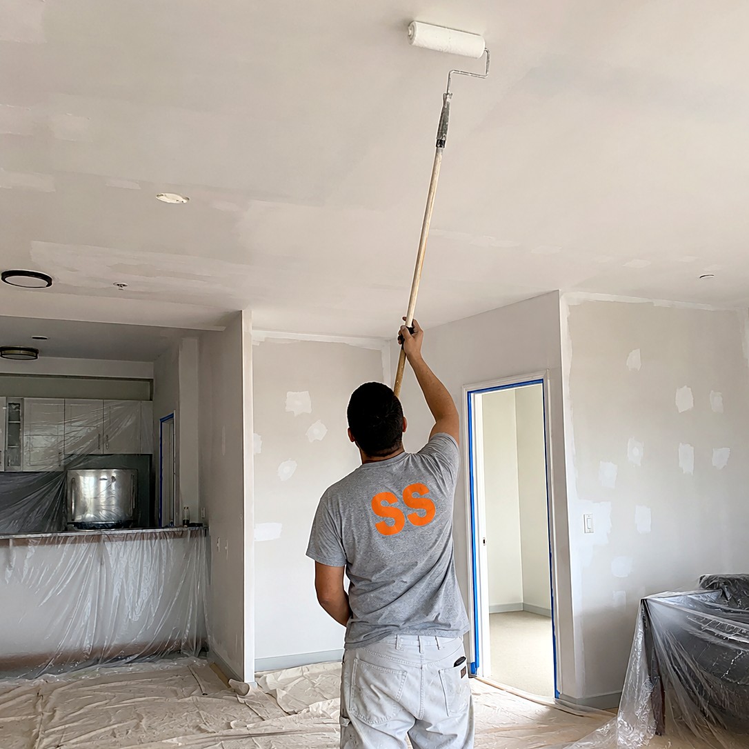 Painter rolling ceiling paint during a whole-apartment repaint with patched walls and taped door frames