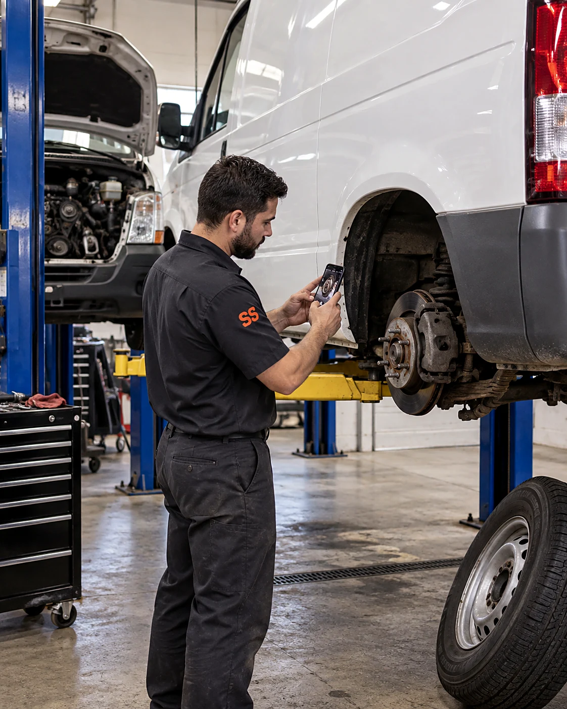 Technician photographing the rear brake rotor of a service van on a lift with the wheel removed, documenting deferred brake work