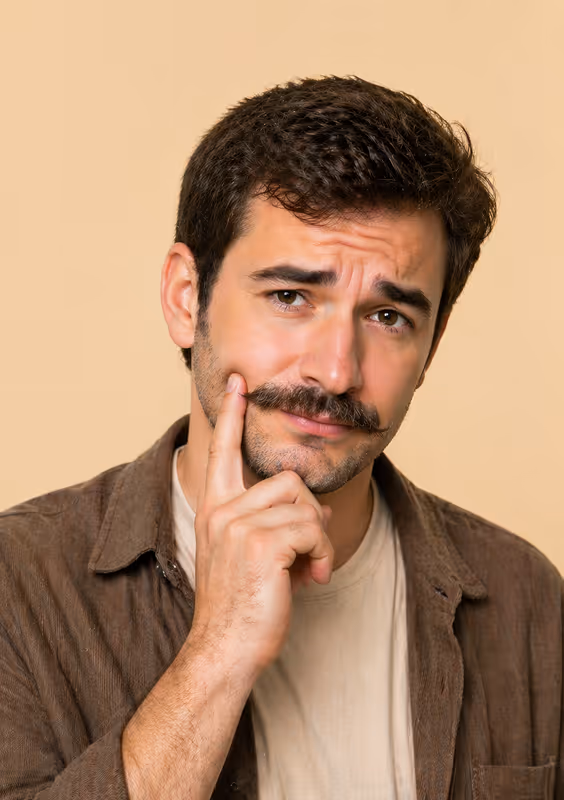 Homme avec une moustache touffue, touchant sa joue, expression pensive sur fond beige.