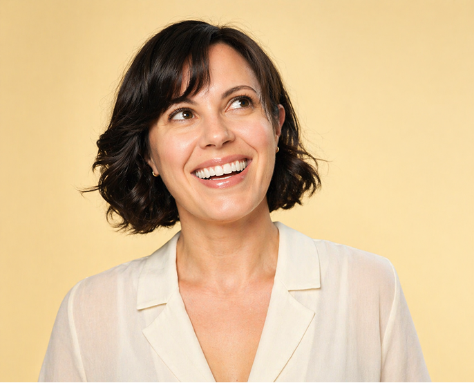 Femme souriante aux cheveux courts et bouclés portant une chemise blanche sur fond jaune.