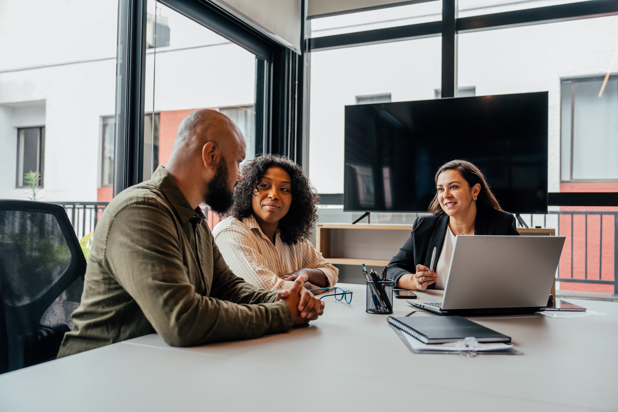 Smiling woman in black jacket talks to a man and woman sitting at a table in a modern office.
