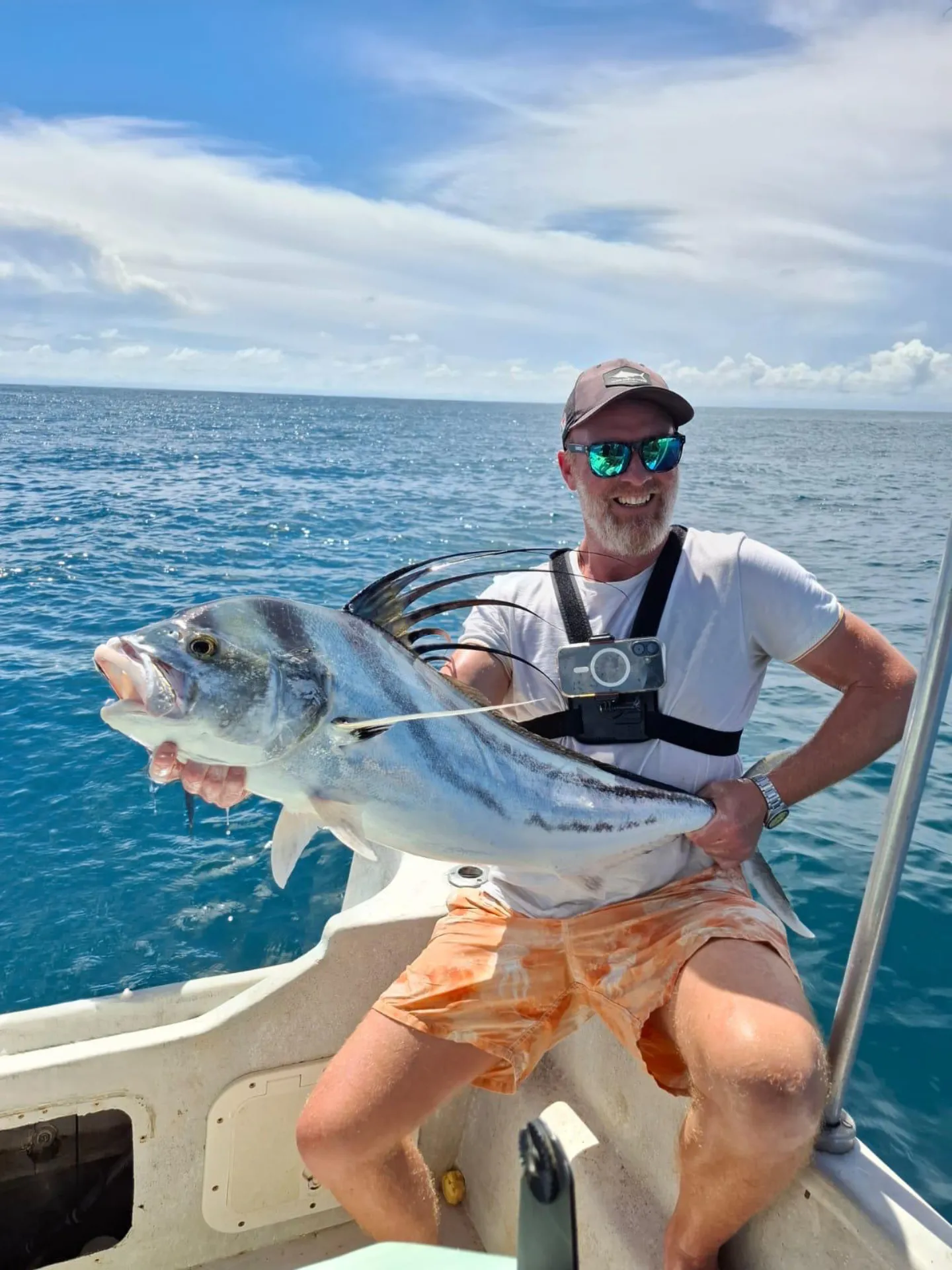 Homme souriant portant des lunettes de soleil et une casquette, tenant un gros poisson sur un bateau avec l'océan en arrière-plan.