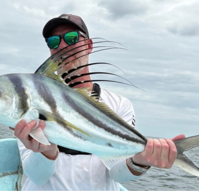 Homme portant des lunettes de soleil et une casquette, tenant un grand poisson-coq sur un bateau en mer sous un ciel nuageux.