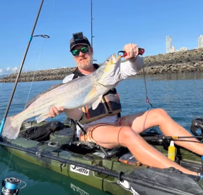 Homme en kayak tenant un gros poisson, avec une canne à pêche et une côte rocheuse en arrière-plan sous un ciel dégagé.