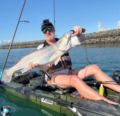 Homme en kayak tenant un gros poisson, avec une canne à pêche et une côte rocheuse en arrière-plan sous un ciel dégagé.