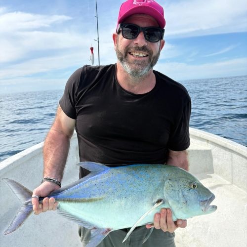 Homme portant une casquette rose et des lunettes de soleil, tenant un grand poisson bleu sur un bateau en mer.