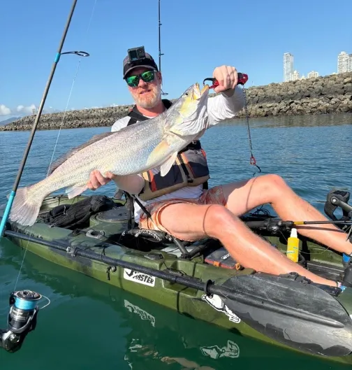 Homme en kayak tenant un gros poisson, avec une canne à pêche et une côte rocheuse en arrière-plan sous un ciel dégagé.