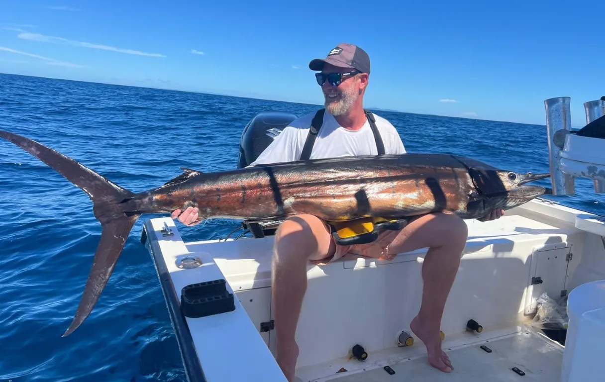 Homme portant des lunettes de soleil et une casquette, tenant un grand marlin sur un bateau dans l'océan.