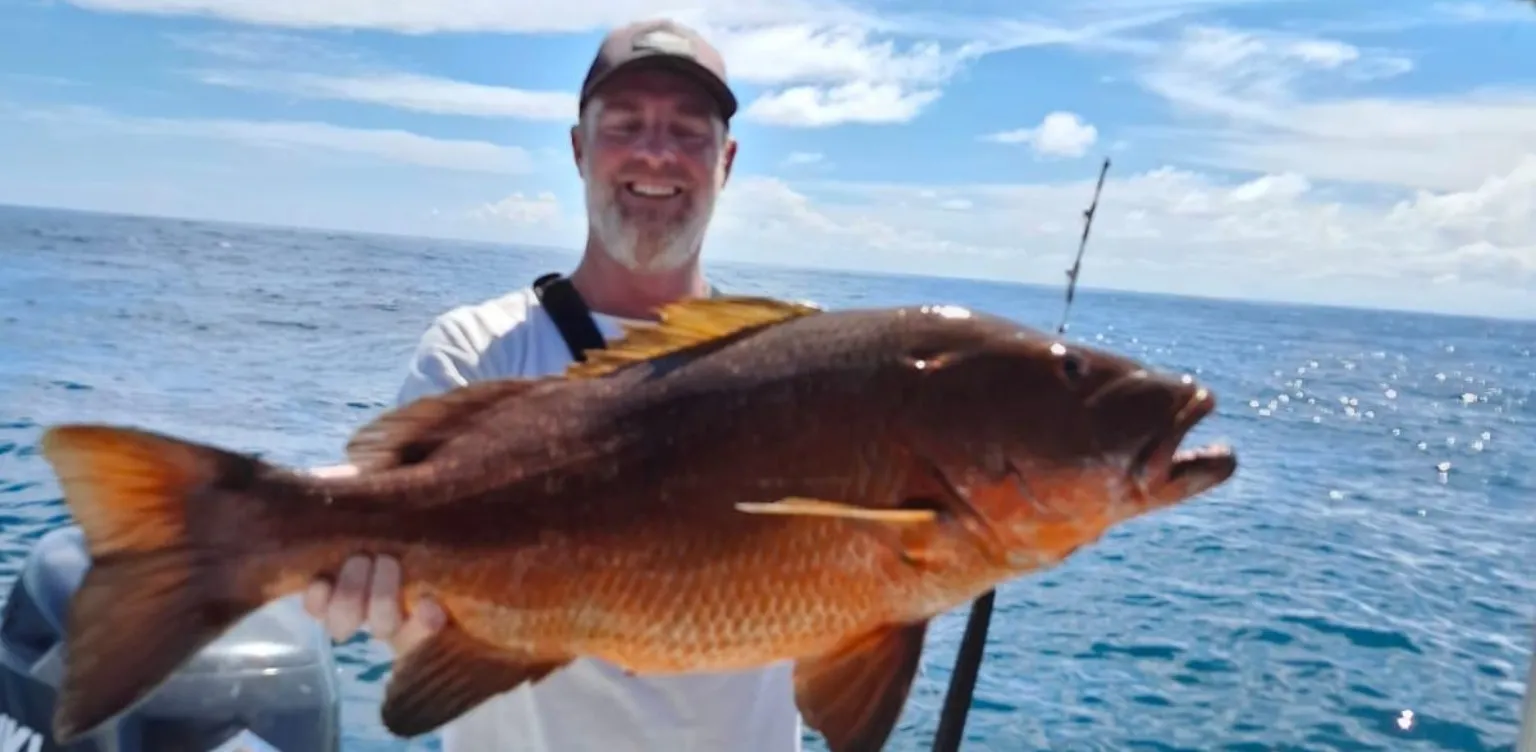 Homme sur un bateau tenant un gros poisson marron sur fond d'océan par une journée ensoleillée.