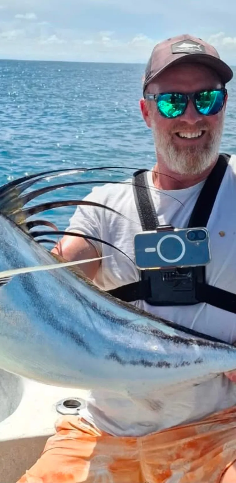 Homme souriant portant des lunettes de soleil et une casquette, tenant un gros poisson sur un bateau avec l'océan en arrière-plan.