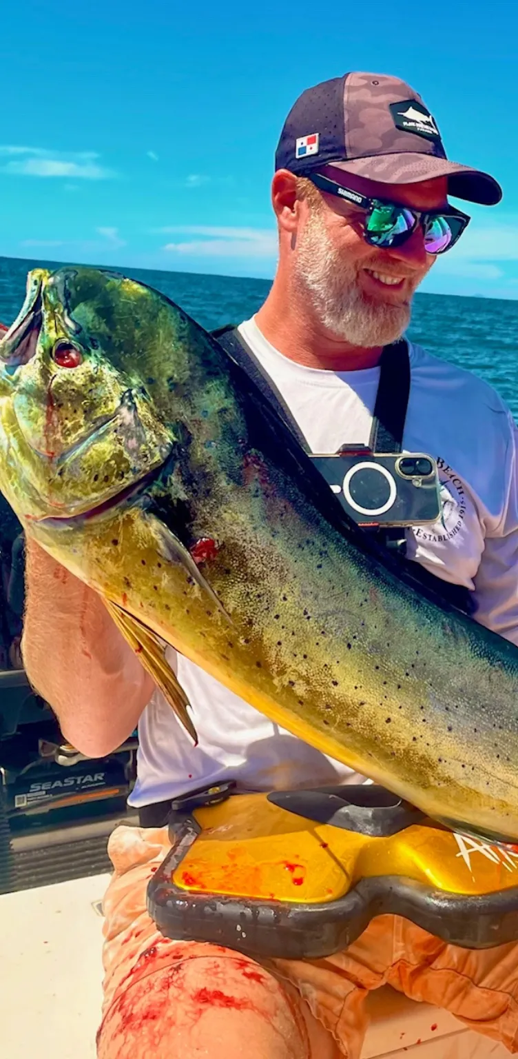 Homme souriant avec un chapeau et des lunettes de soleil, tenant un grand poisson aux couleurs vives sur un bateau dans l'océan.