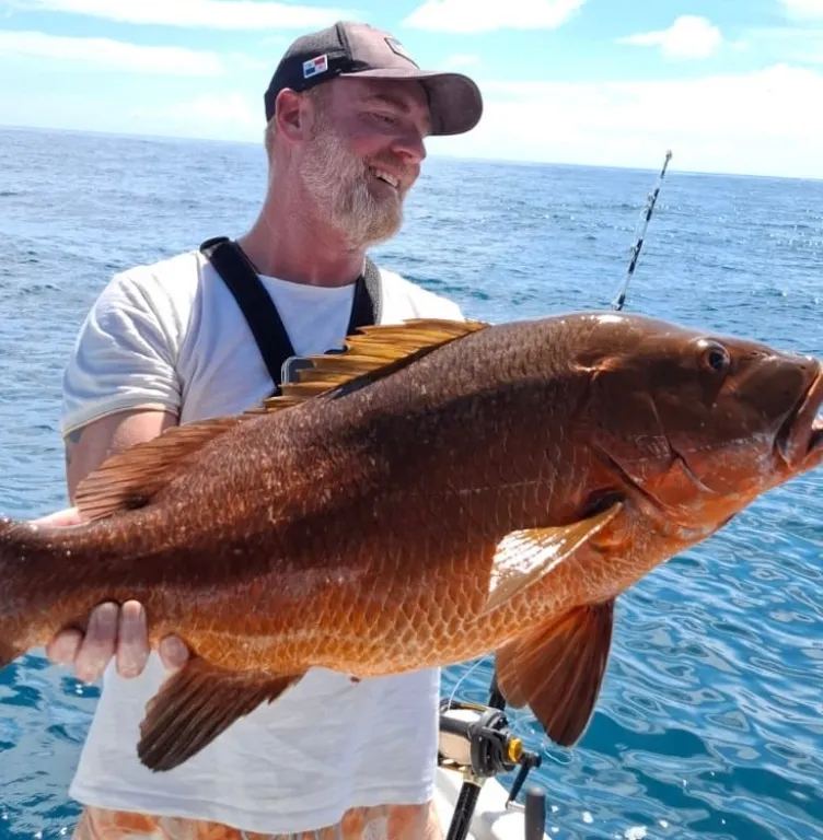 Homme souriant portant une casquette, tenant un gros poisson marron sur un bateau avec l'océan en arrière-plan.