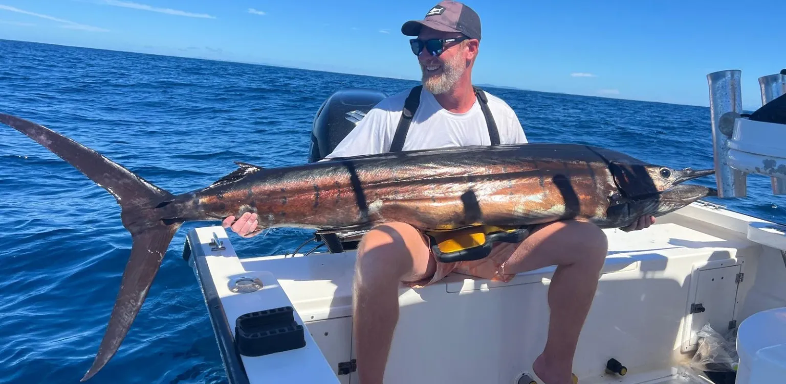 Homme sur un bateau tenant un gros poisson avec l'océan bleu et le ciel en arrière-plan.