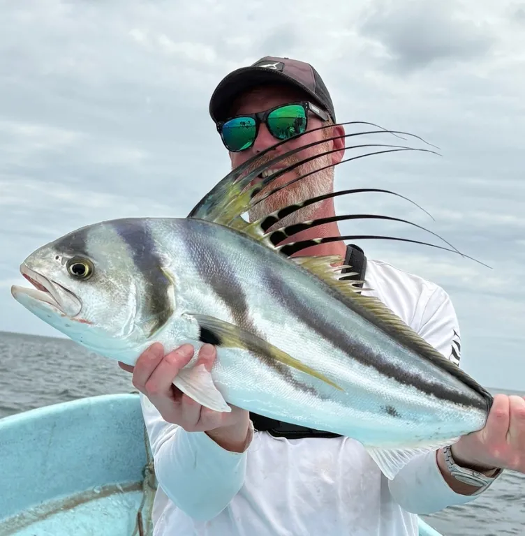 Homme portant des lunettes de soleil et une casquette, tenant un grand poisson-coq sur un bateau en mer sous un ciel nuageux.