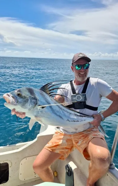 Homme souriant portant des lunettes de soleil et une casquette, tenant un gros poisson sur un bateau avec l'océan et le ciel en arrière-plan.