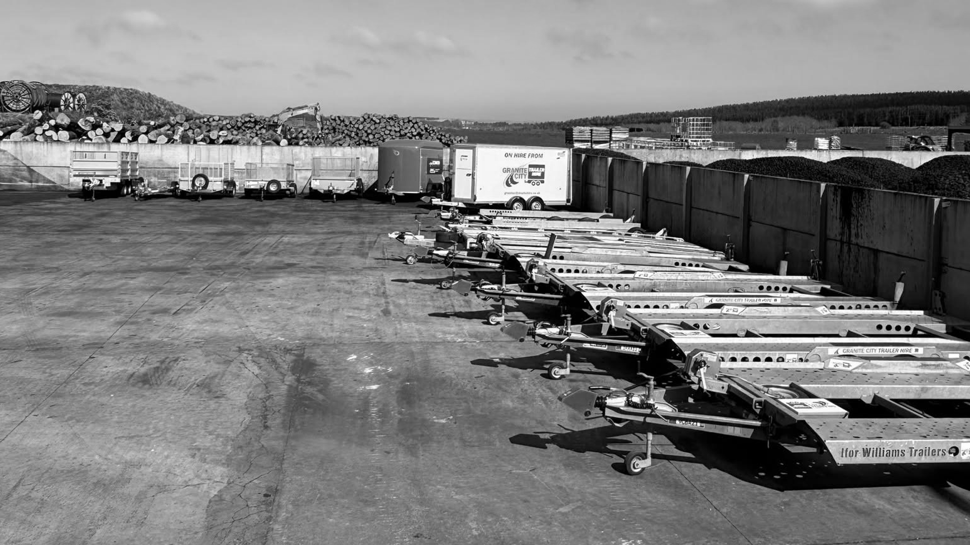 Rows of trailers and trailer frames parked in a large outdoor industrial yard with stacked logs and materials.