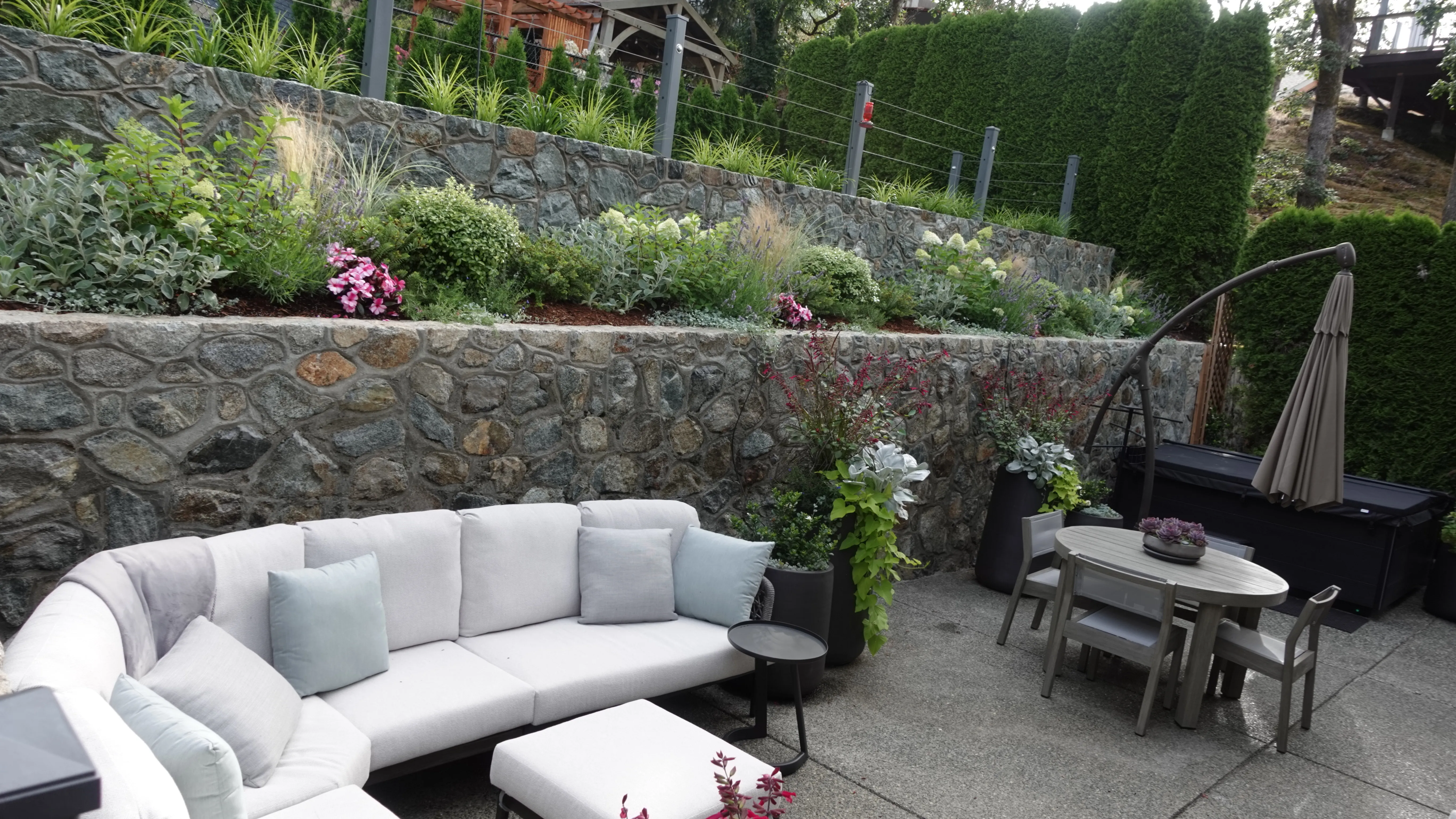 Stone-walled backyard patio with sectional seating, statement planters and terraced garden beds at the Strawberry Vale Residence, Victoria BC