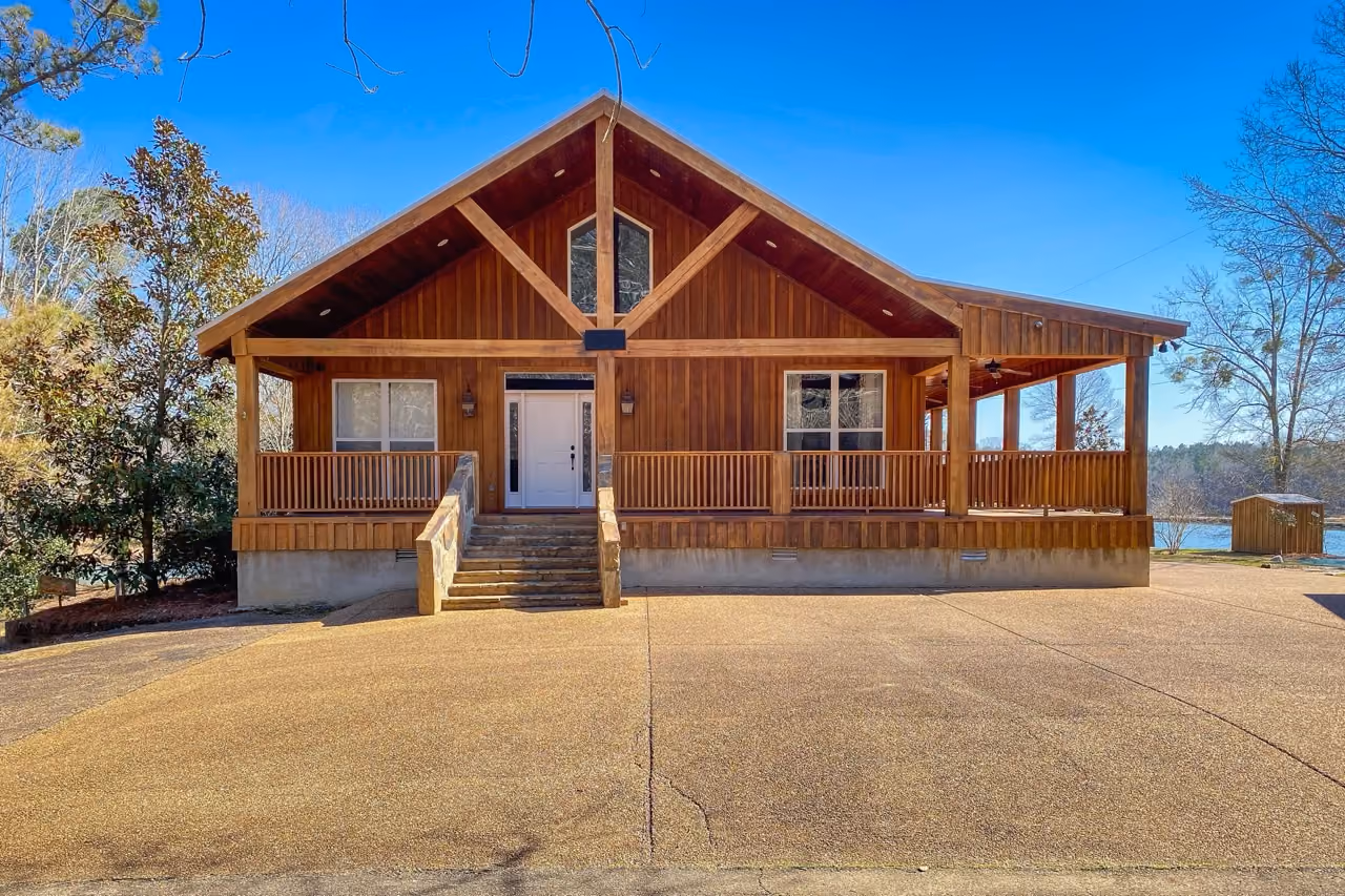 Front view of a brown wooden house for sale in central Mississippi with a covered porch, white front door, and steps leading to the entrance, set against a clear blue sky.