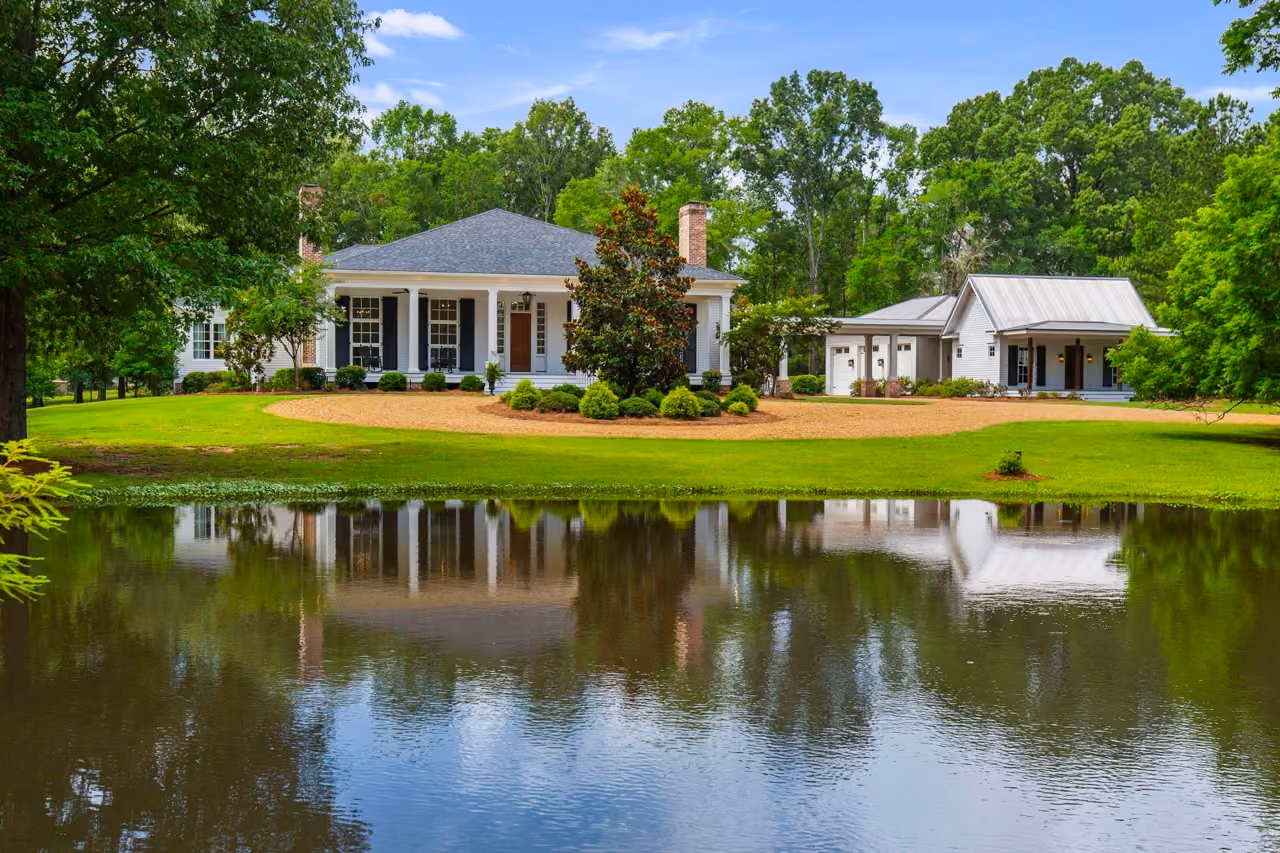 A white traditional house for sale in central Mississippi with columns and black shutters beside a smaller white building with a metal roof, both reflected in a large pond surrounded by green grass and trees.