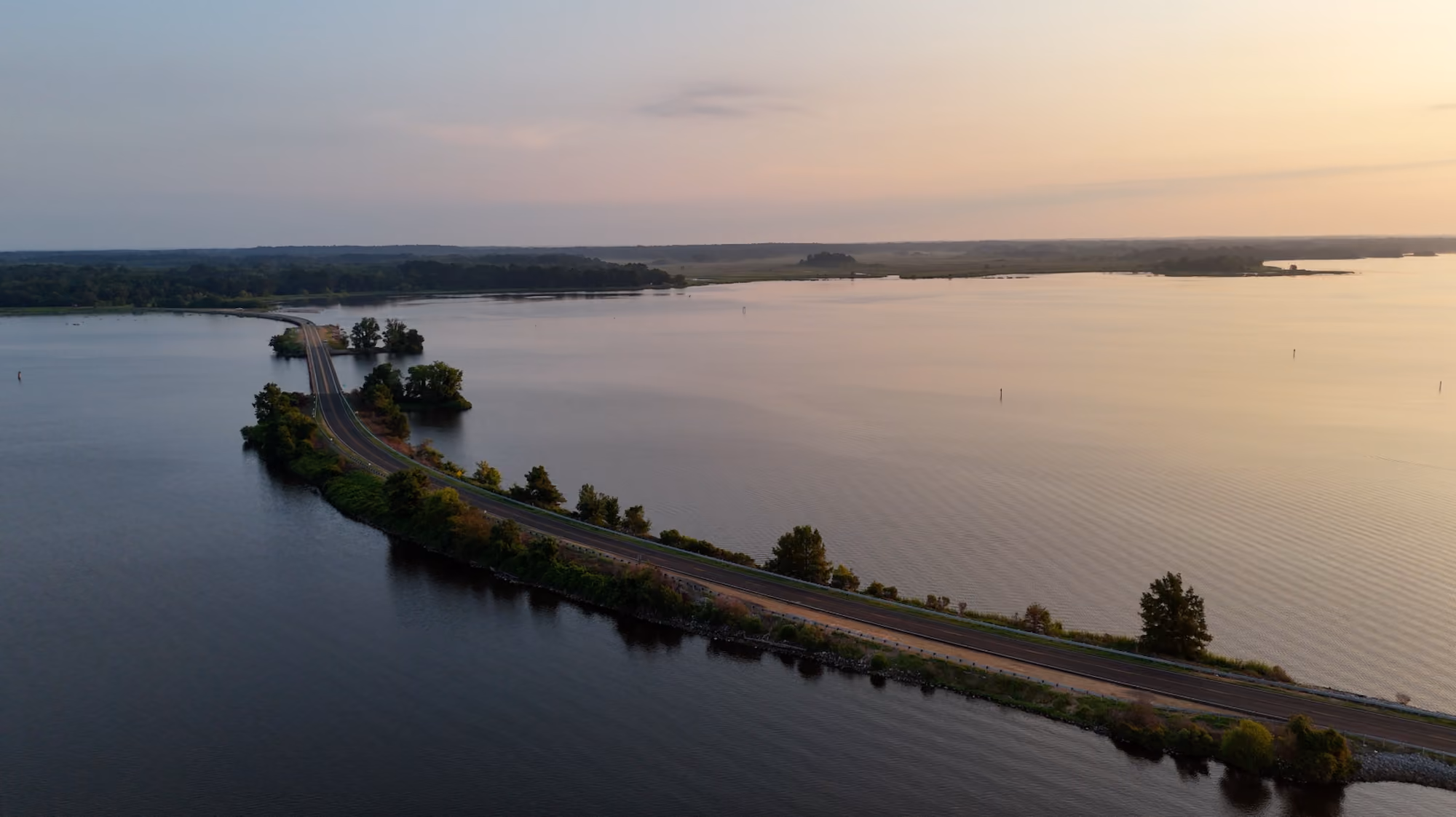 A winding road in central Mississippi cuts across a narrow strip of land surrounded by calm water under a clear sky at sunset.