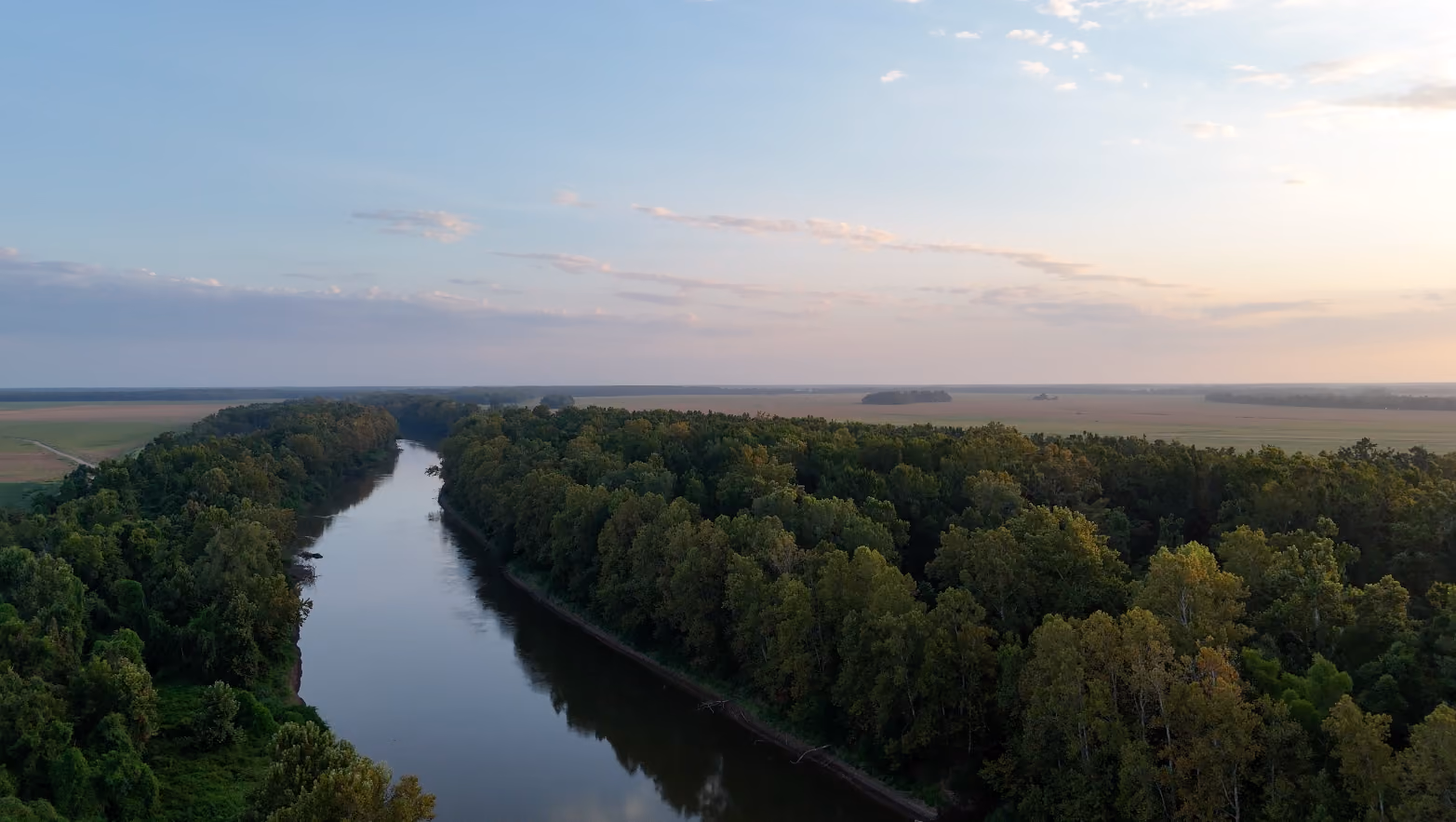 Aerial view of central Mississippi  river curving through dense green forest with open fields in the distance under a partly cloudy sky at sunrise or sunset.