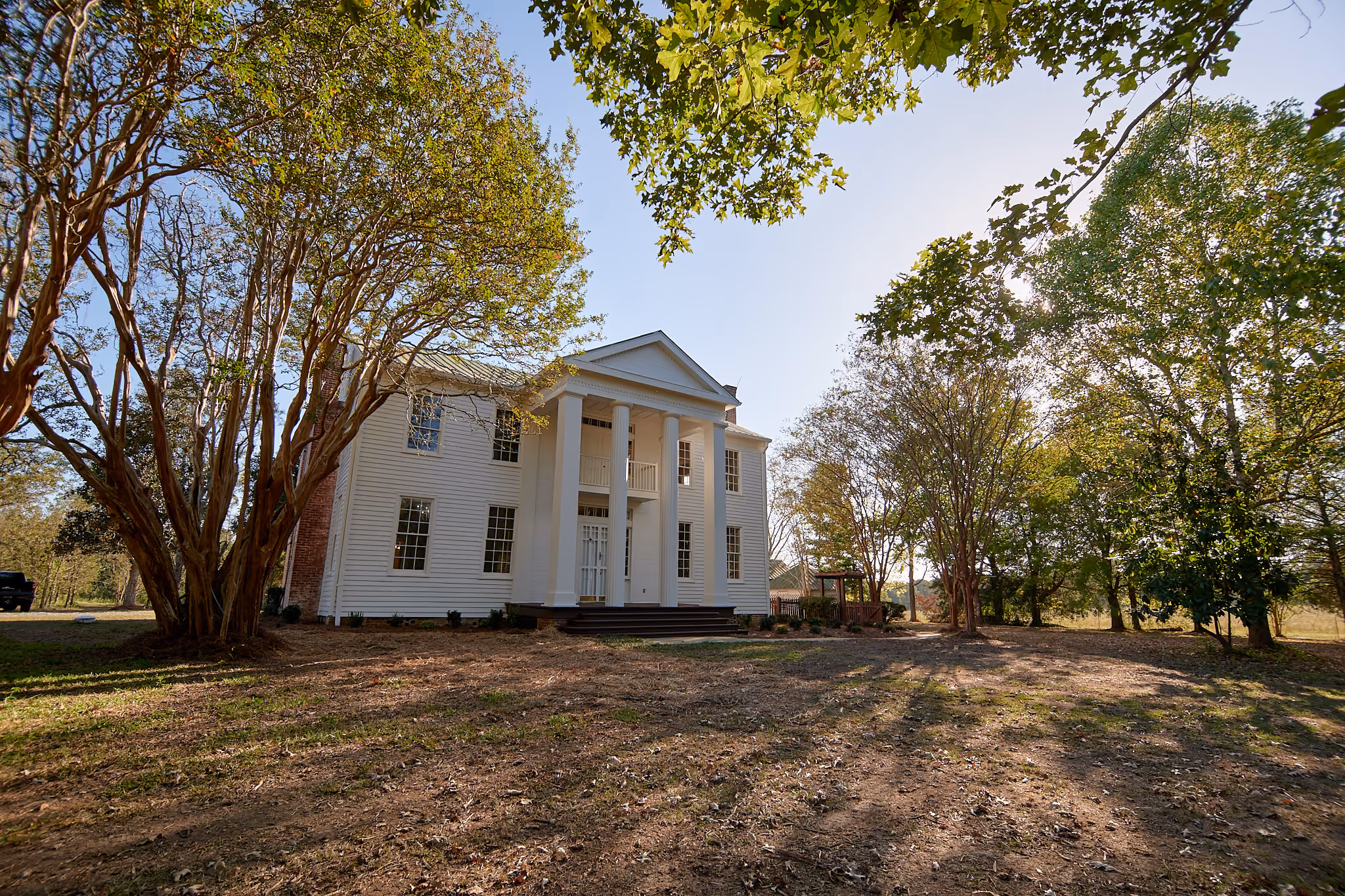 Two-story white colonial-style house for sale in central Mississippi with tall columns at the entrance, surrounded by large trees under a clear sky.