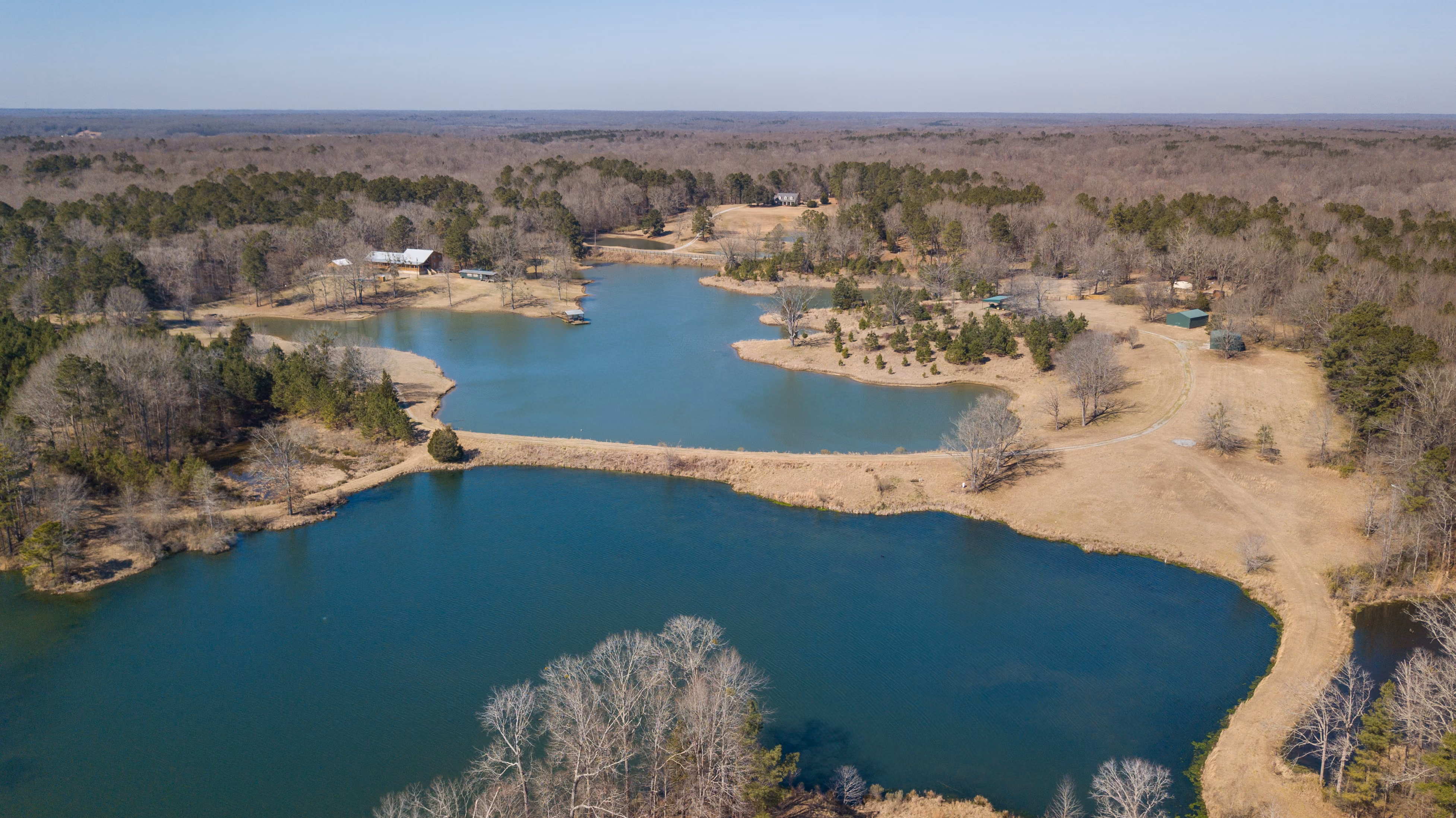 Aerial view of a rural landscape in central Mississippi featuring multiple ponds bordered by dry grass and scattered leafless trees, with a mix of dense forest and open clearings extending to the horizon under a clear sky.