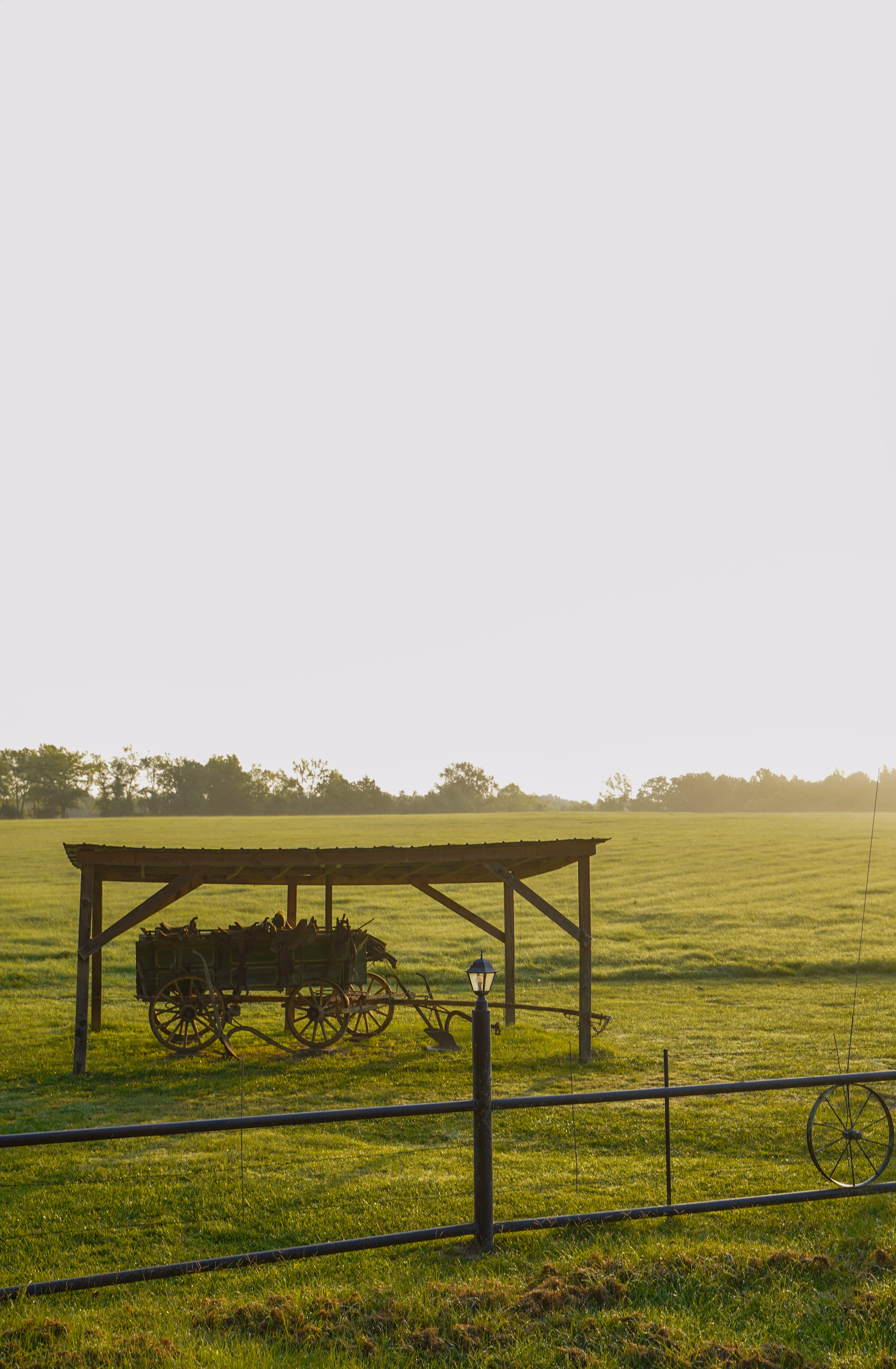 A wooden covered wagon with wooden wheels parked in a grassy field during sunset, with a black fence and lamp post in the foreground and trees in the distance.