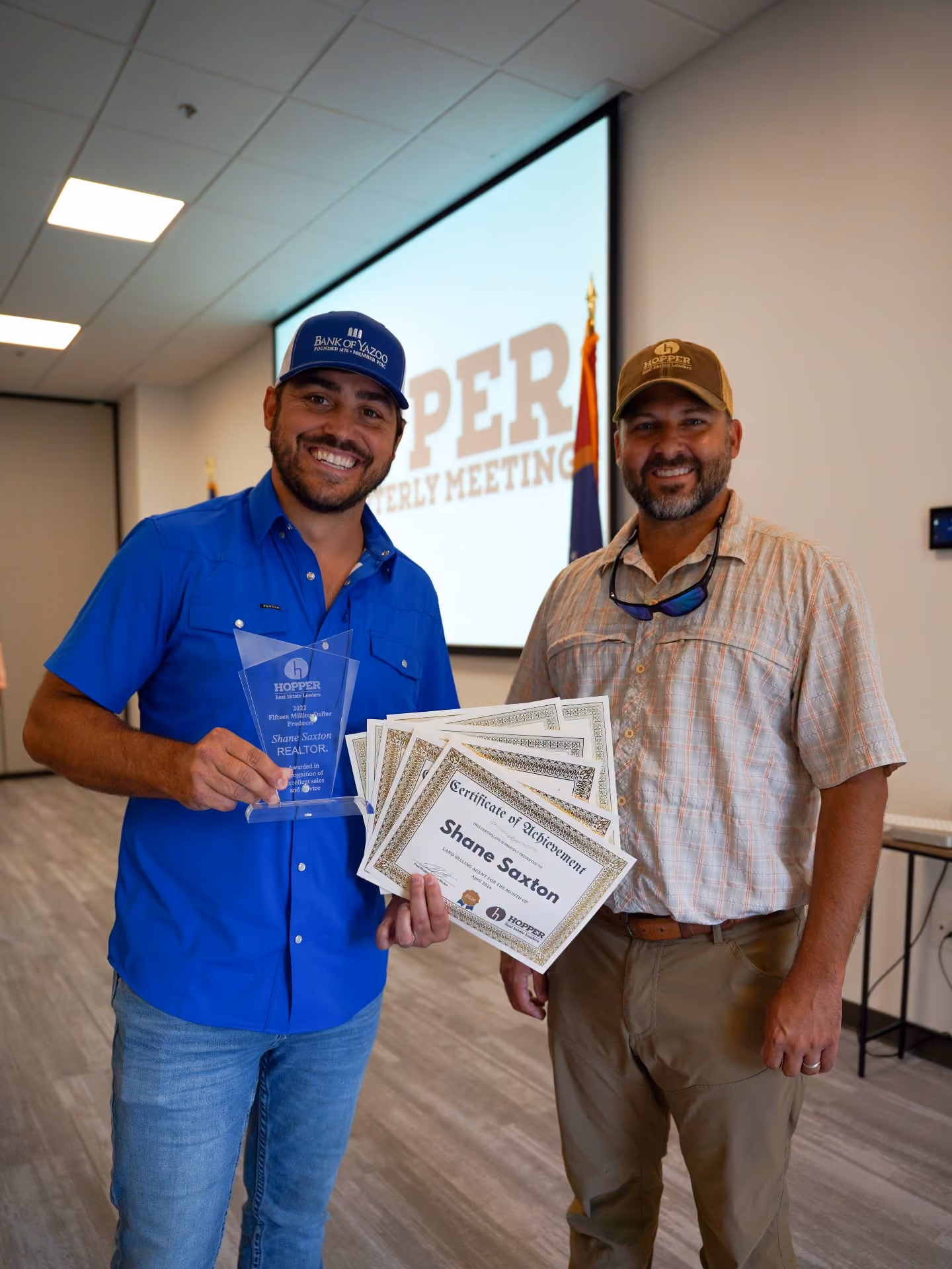 Two Hopper Real Estate Leaders standing indoors at a meeting room, one agent in a blue shirt and cap holding a trophy and several certificates, the certificates read 'Certificate of Achievement Shane Saxton.' The other man, Paul Hopper, wears a light plaid shirt and brown pants smiling beside him.