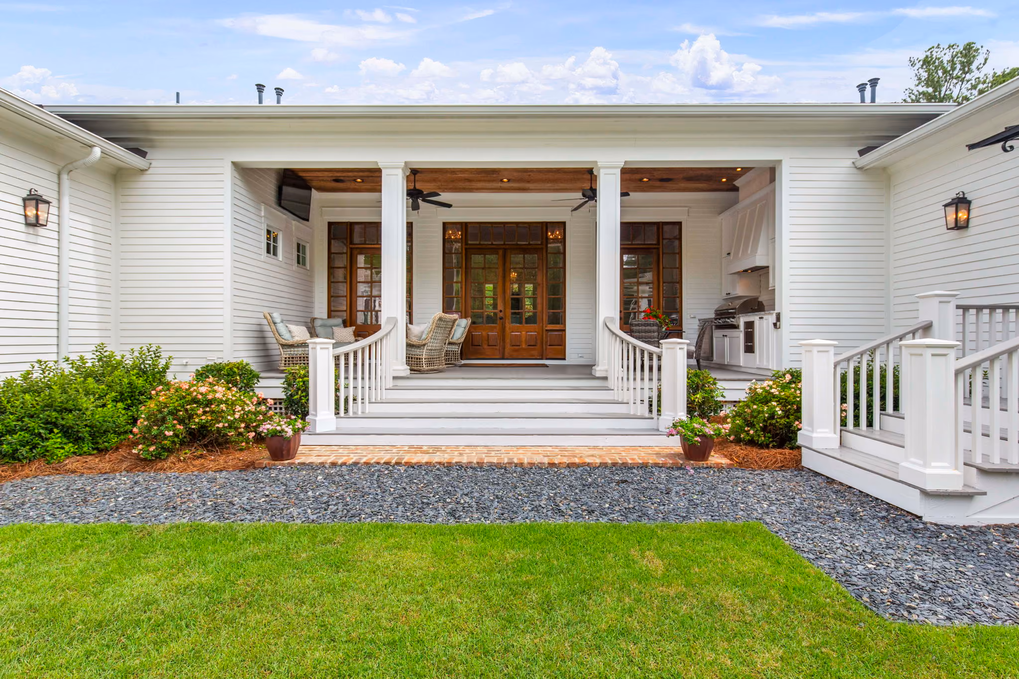 Home in central Mississippi with porch featuring three columns, wooden staircase with railings, wicker chairs, and double wooden doors, surrounded by green lawn and flower bushes.