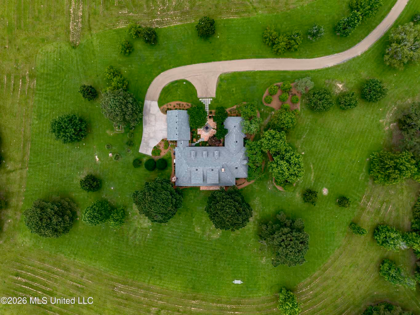 Aerial view of a single large house for sale in Central Mississippi with a gray roof surrounded by green lawn and scattered trees, with a curved driveway leading to the house.