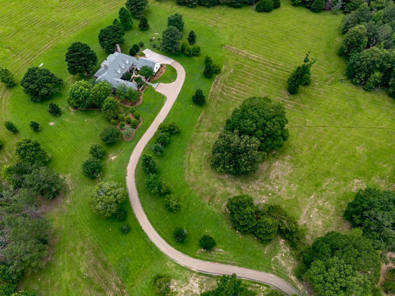 Aerial view of a large Central Mississippi house with a gray roof surrounded by green lawn and trees, accessed by a curved paved driveway through spacious grassy land.