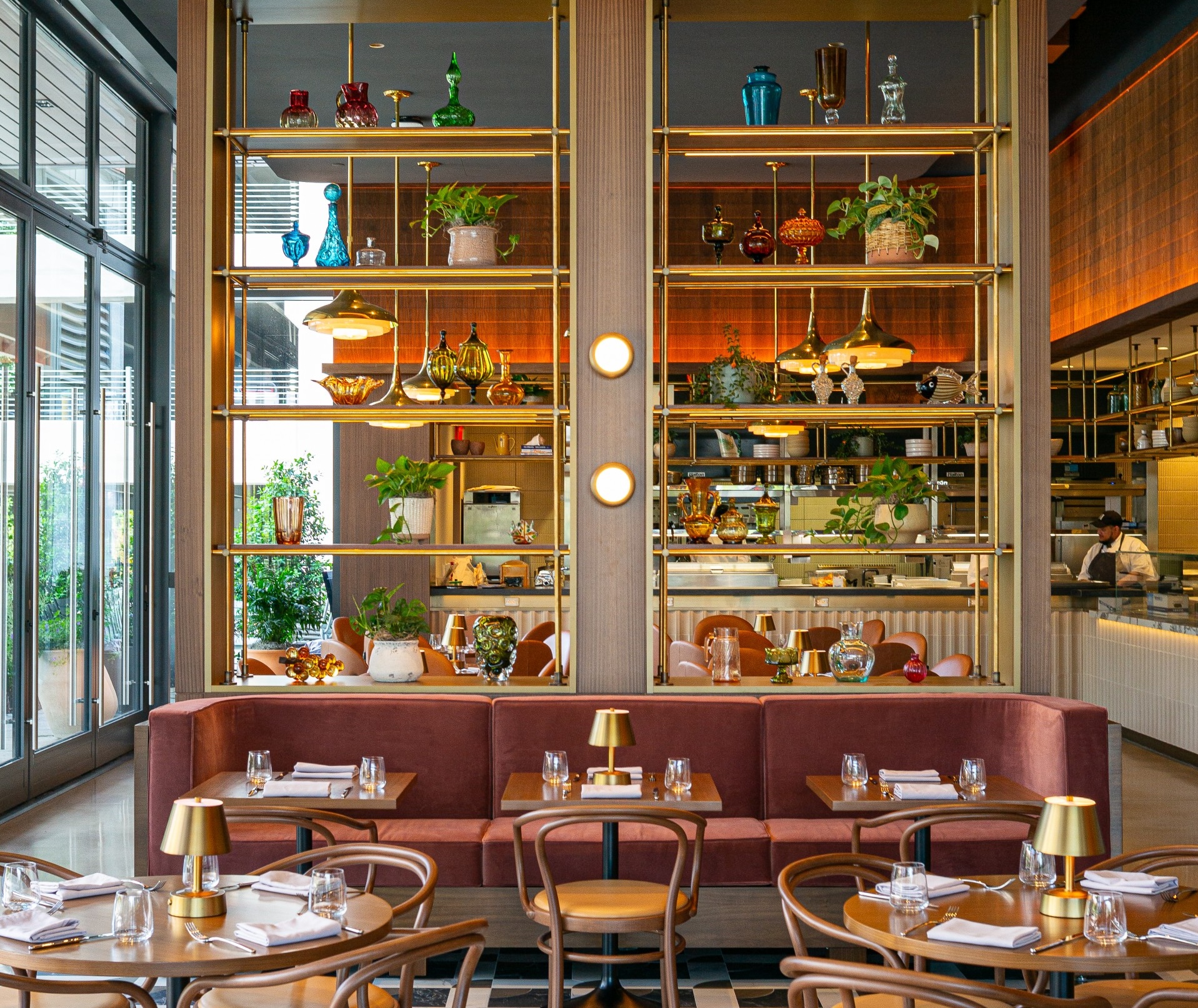 West Wing dining area at The Morris Proper with a pink banquette, brass lamps, and floor-to-ceiling shelves of plants and colored glass