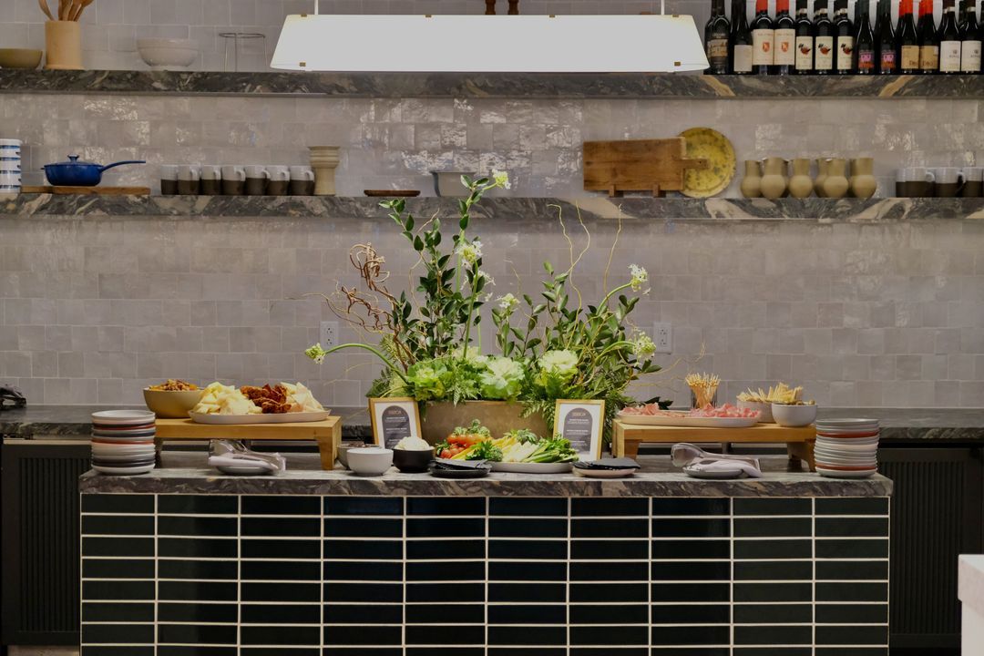 Grazing station with cheese, charcuterie, and crudités around a tall floral arrangement in front of the open kitchen at The Morris Proper
