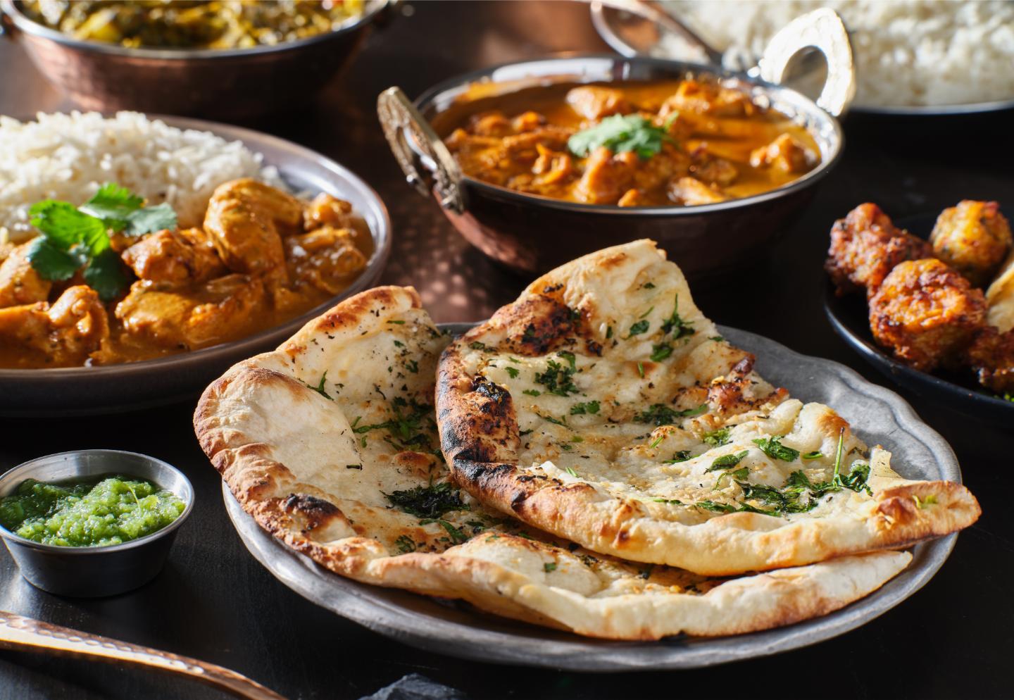 Plate of naan bread garnished with herbs, surrounded by dishes of curry, rice, and green chutney.
