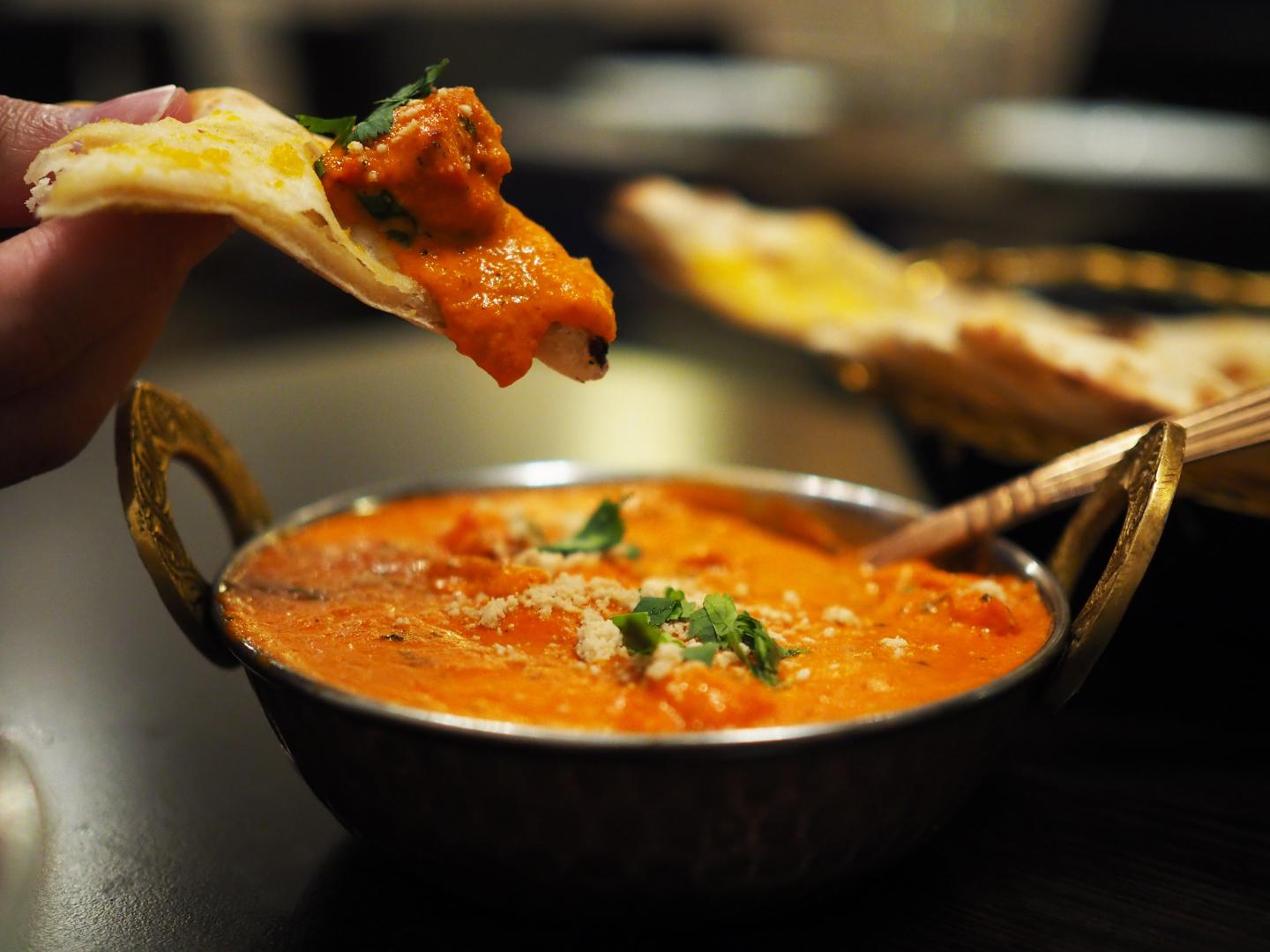 Hand dipping naan bread into a bowl of creamy orange curry garnished with herbs.